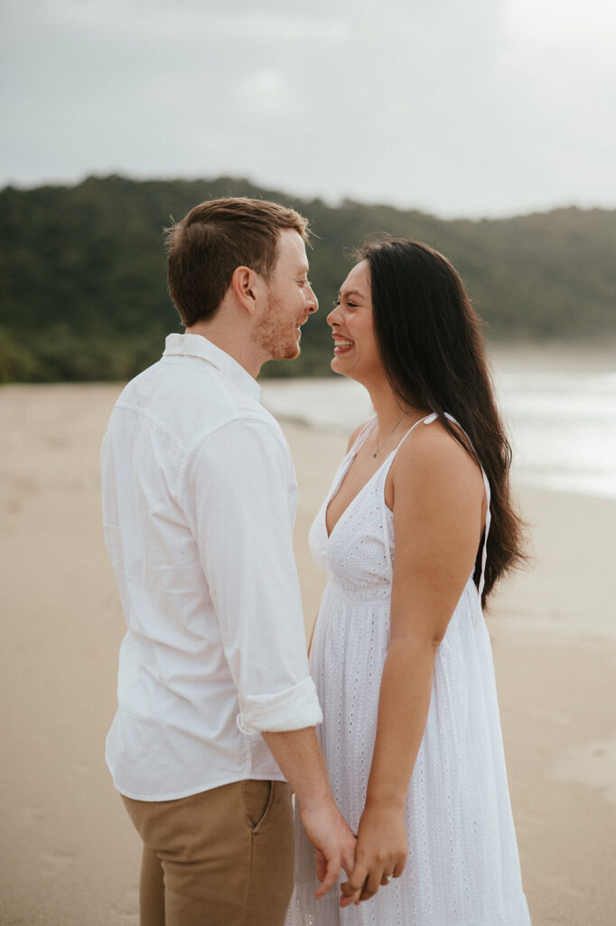 Andrea and Ross holding hands while walking barefoot along the shoreline with soft waves behind them.
