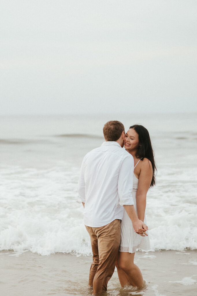 Andrea and Ross holding hands while walking barefoot along the shoreline with soft waves behind them.
