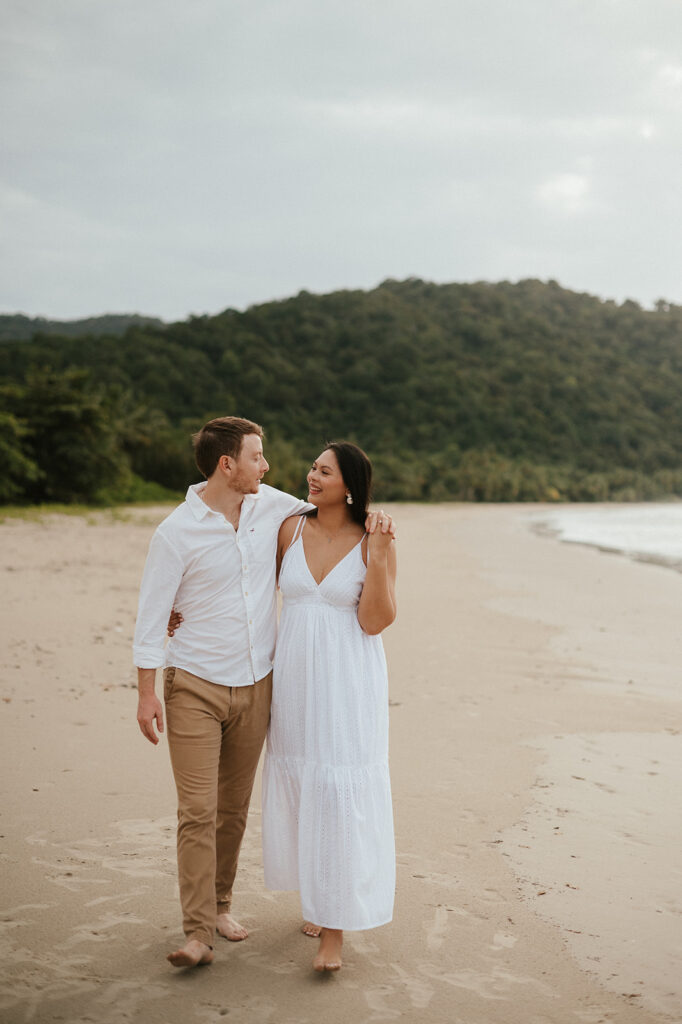Andrea and Ross holding hands while walking barefoot along the shoreline with soft waves behind them.

