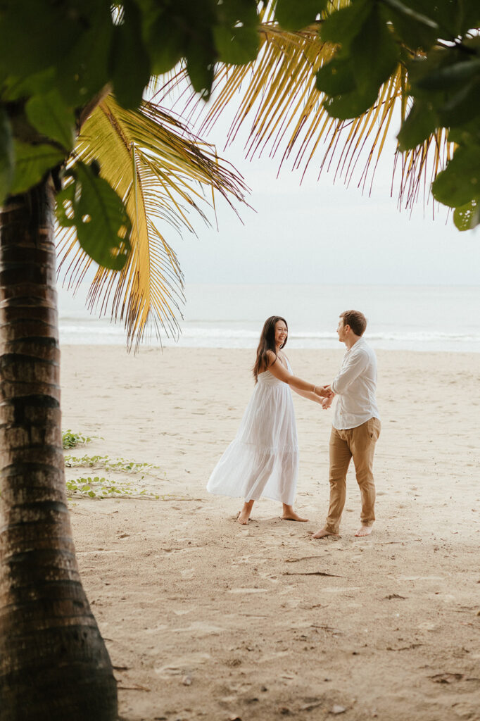 Andrea and Ross holding hands while walking barefoot along the shoreline with soft waves behind them.
