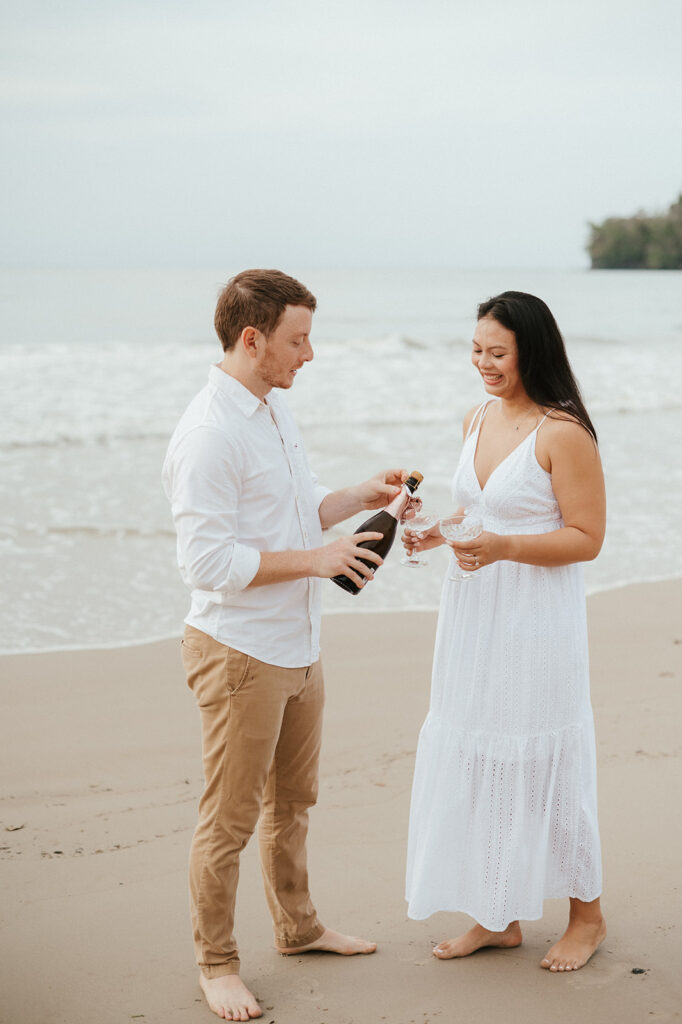 Newly engaged couple laughing and spraying champagne during a celebration on the sand at Las Cuevas Beach.
