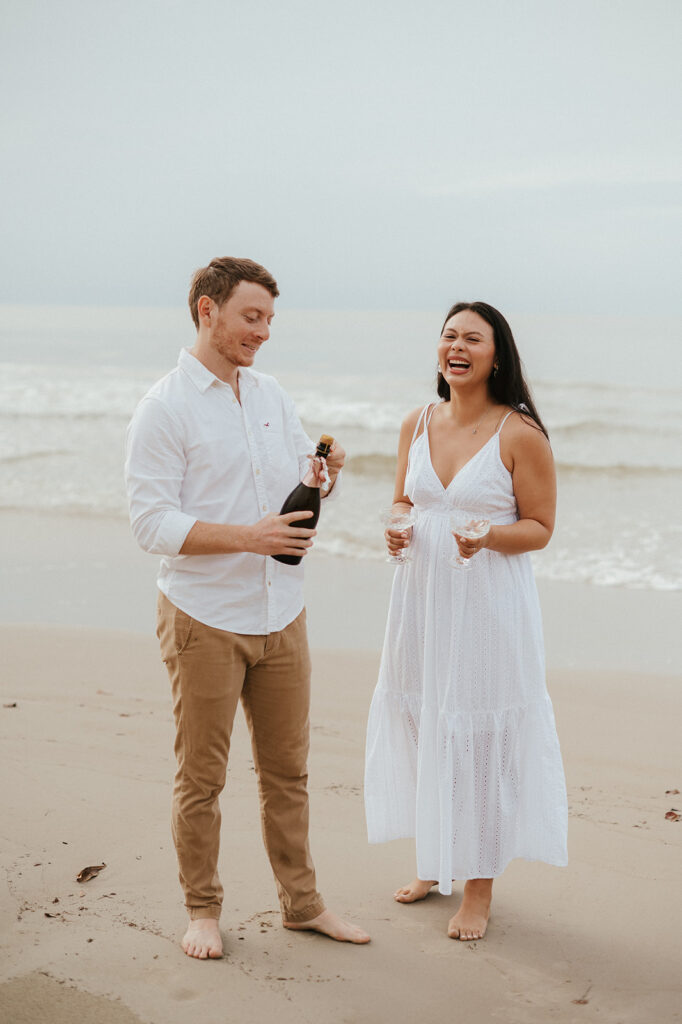 Newly engaged couple laughing and spraying champagne during a celebration on the sand at Las Cuevas Beach.
