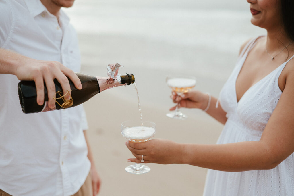Newly engaged couple laughing and spraying champagne during a celebration on the sand at Las Cuevas Beach.
