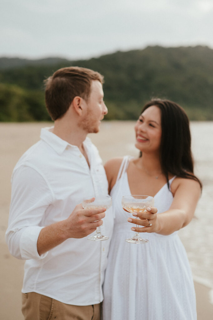 Andrea and Ross holding hands while walking barefoot along the shoreline with soft waves behind them.
