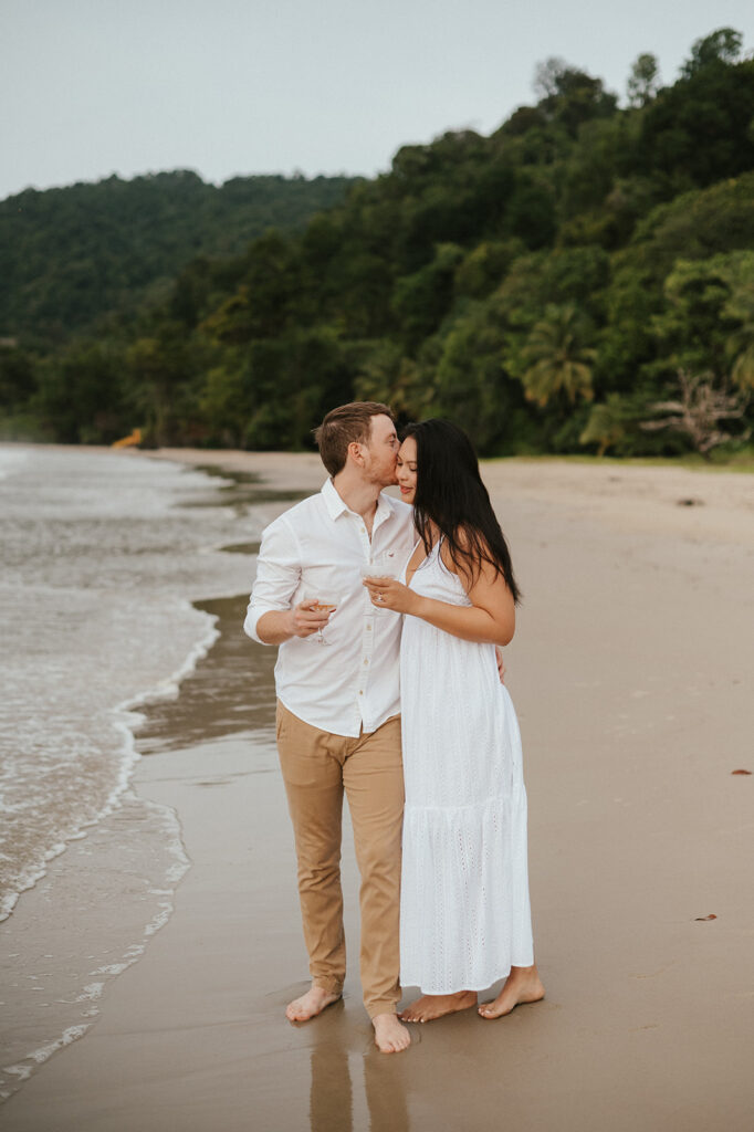 Andrea and Ross holding hands while walking barefoot along the shoreline with soft waves behind them.

