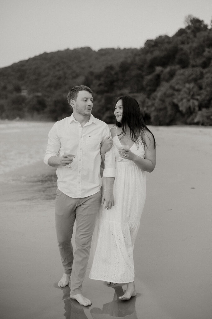 Andrea and Ross holding hands while walking barefoot along the shoreline with soft waves behind them.
