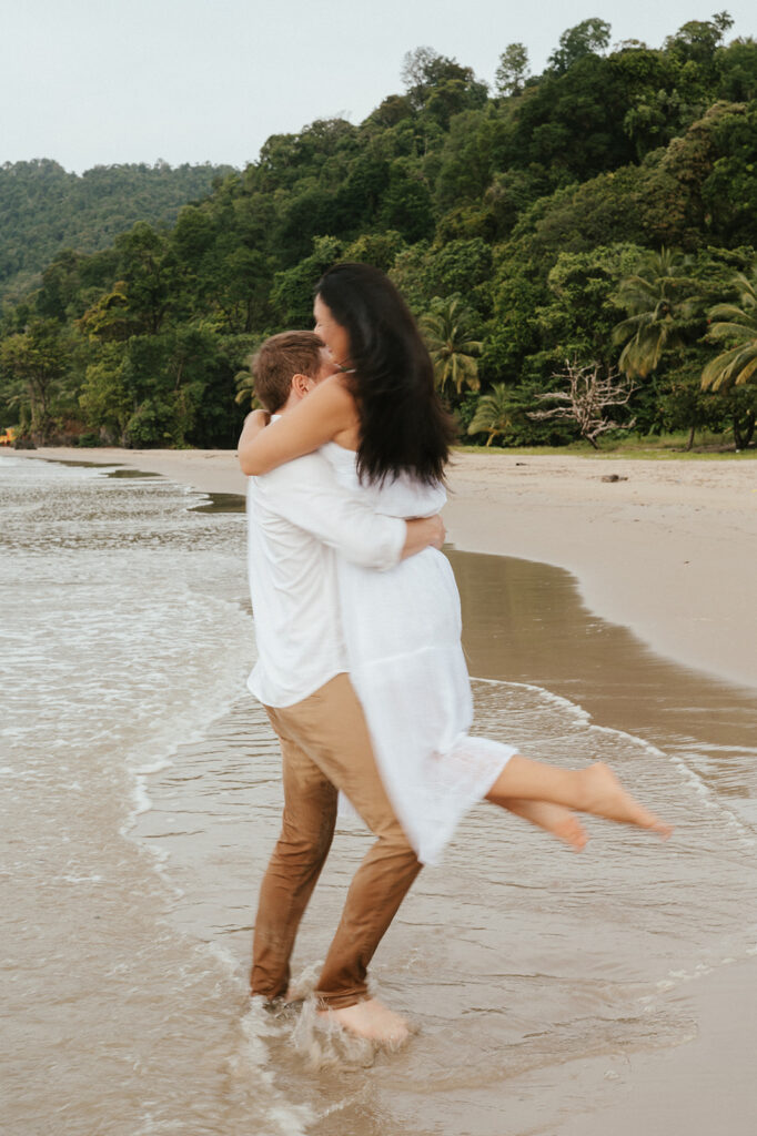 Andrea and Ross holding hands while walking barefoot along the shoreline with soft waves behind them.
