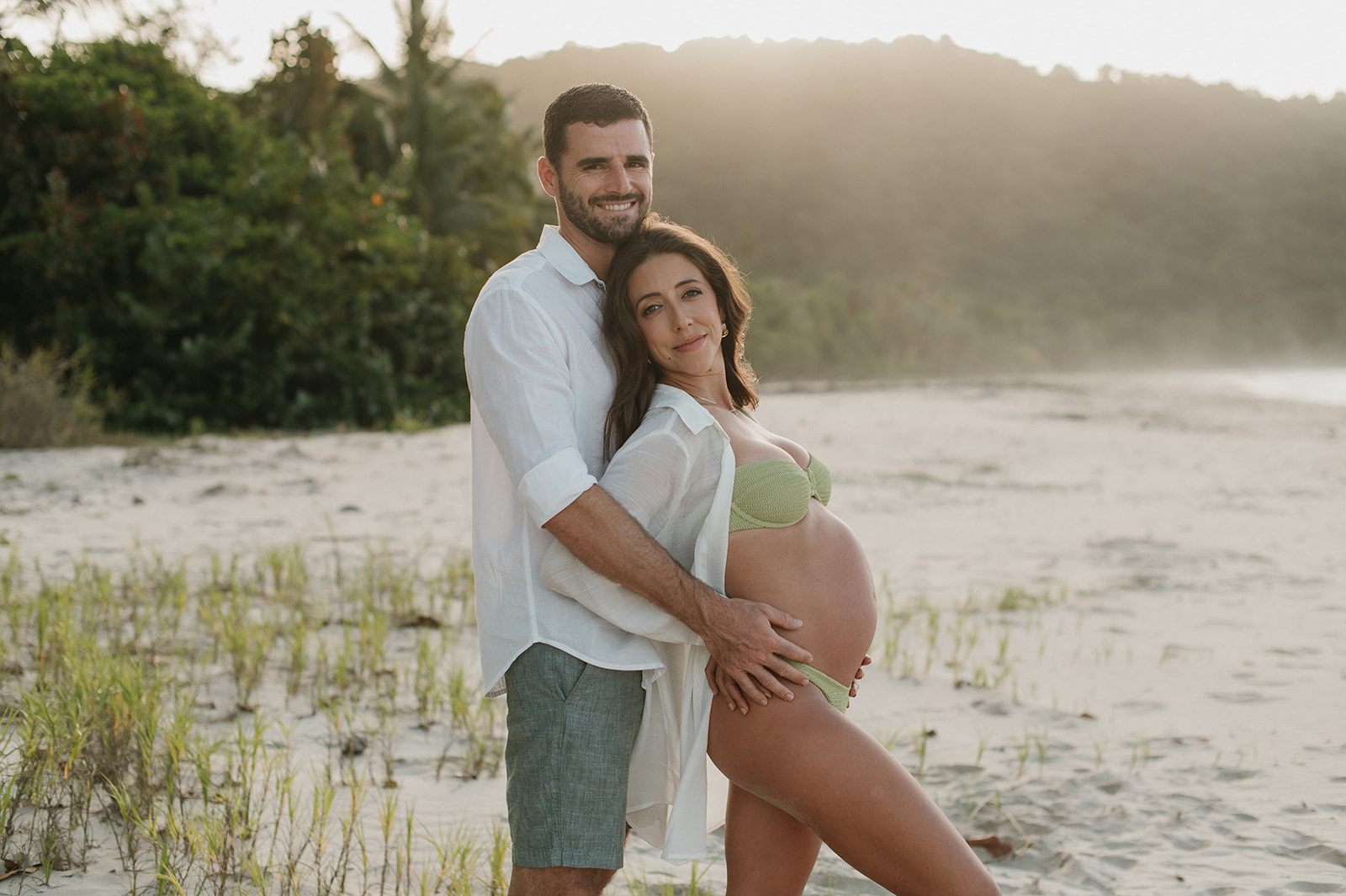 A pregnant woman in a bikini leans back against her husband, during golden hour at a Trinidad beach